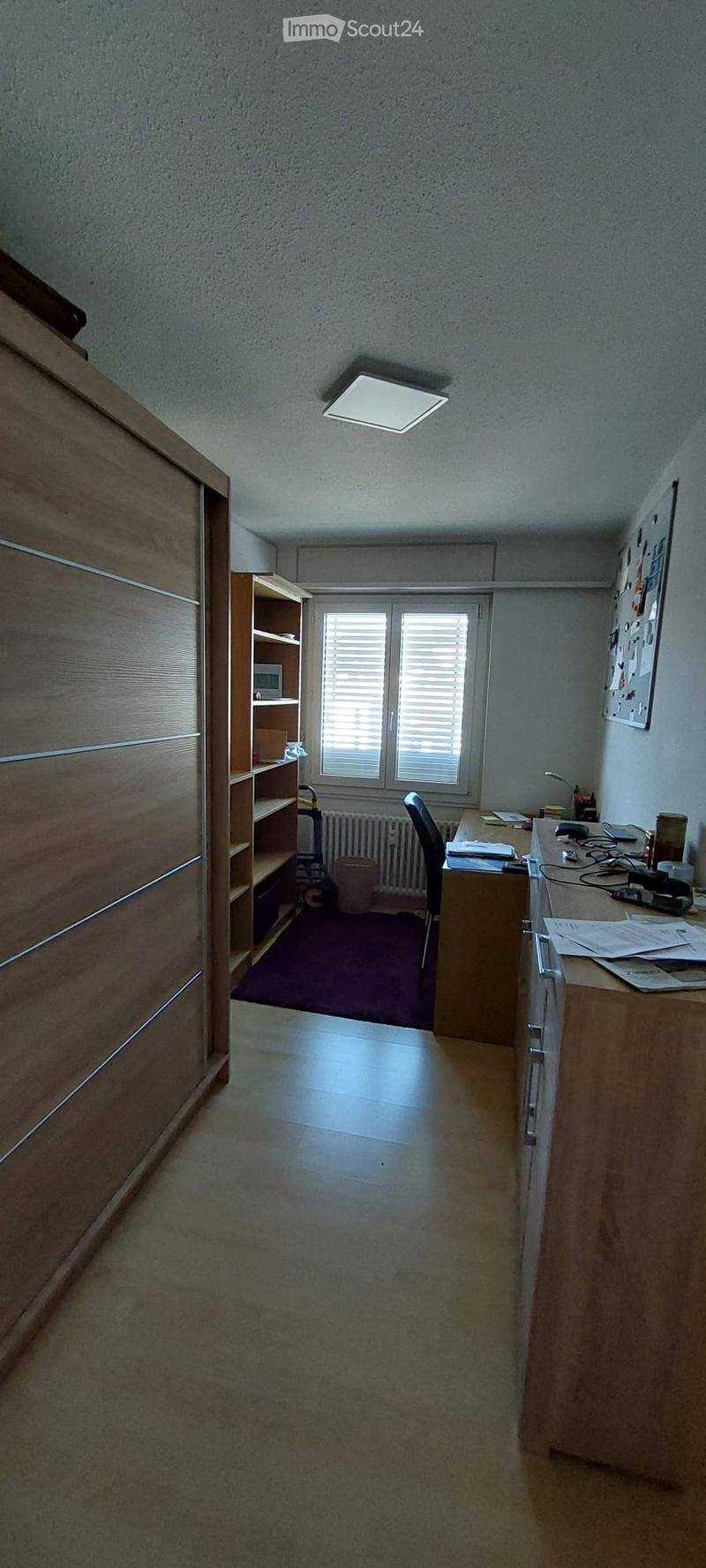 Office space with a wooden desk, chair, laptop, bookshelf, radiator, and trash bin. The room has a purple carpeted area and a large window with blinds.
