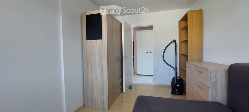 A modern, furnished bedroom with a bed, wardrobe, shelf, vacuum cleaner, and desk.