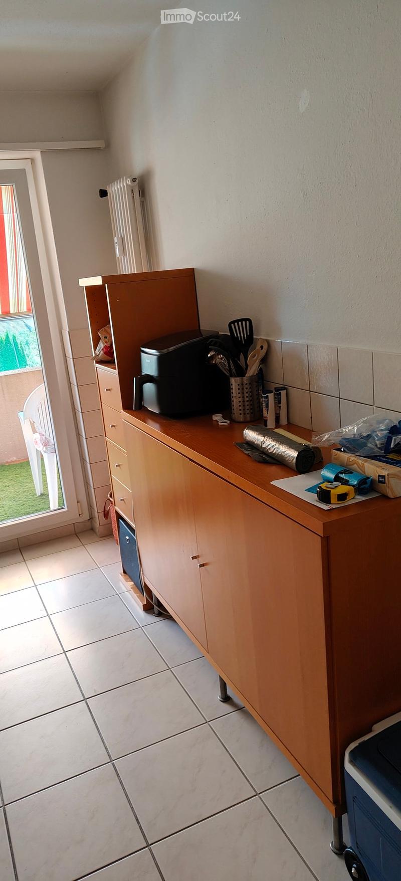 A small kitchen with light wooden cabinets, a tiled floor, and a countertop holding a printer, utensils, and other items. A white chair is visible near the window.