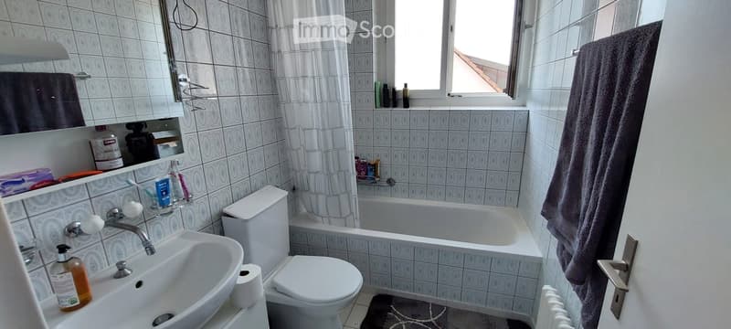 Bathroom with white tiles, a sink, toilet, bathtub, mirror, and towel. Personal care items are visible on the sink and shelf.