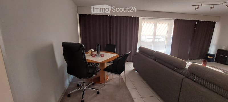 A modern living and dining area with a sofa, dining table, and chairs. The room features large windows with curtains and a tiled floor.