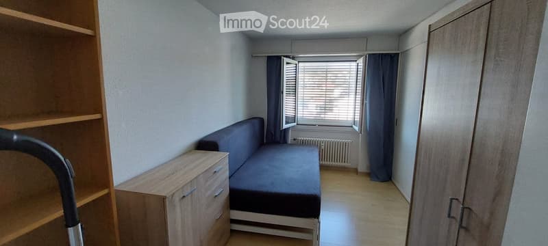 A small, furnished room with a blue sofa, a bed, a wooden cabinet, and a shelf. The room has light-colored walls and a window with blinds.