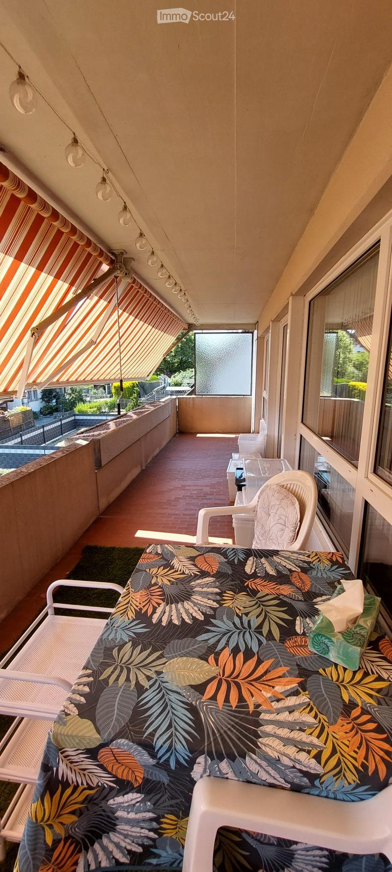 Balcony with a table covered in a leaf-patterned cloth, two white plastic chairs, and a small green rug.