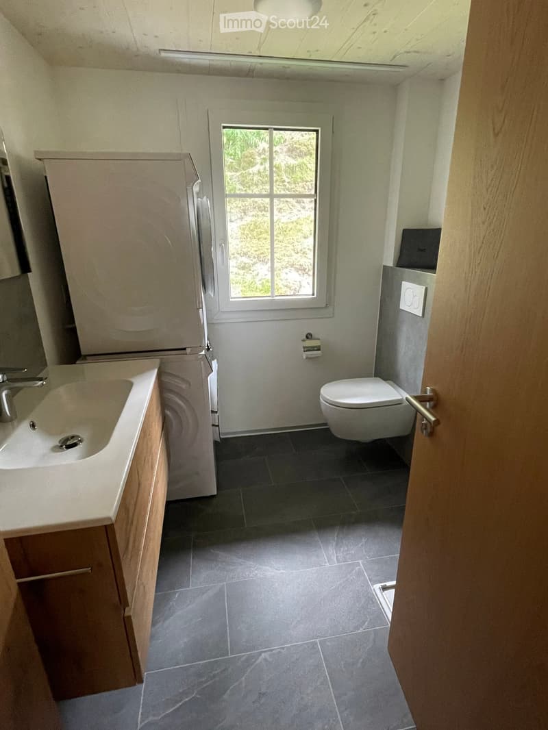 Modern bathroom with a sink, toilet, and wooden cabinet. Features dark tiled flooring and a window with a view of greenery.