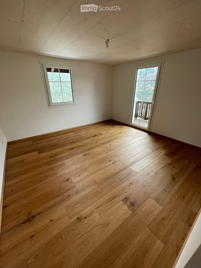 Empty room with wooden flooring, white walls, and two windows, one of which opens to a balcony.