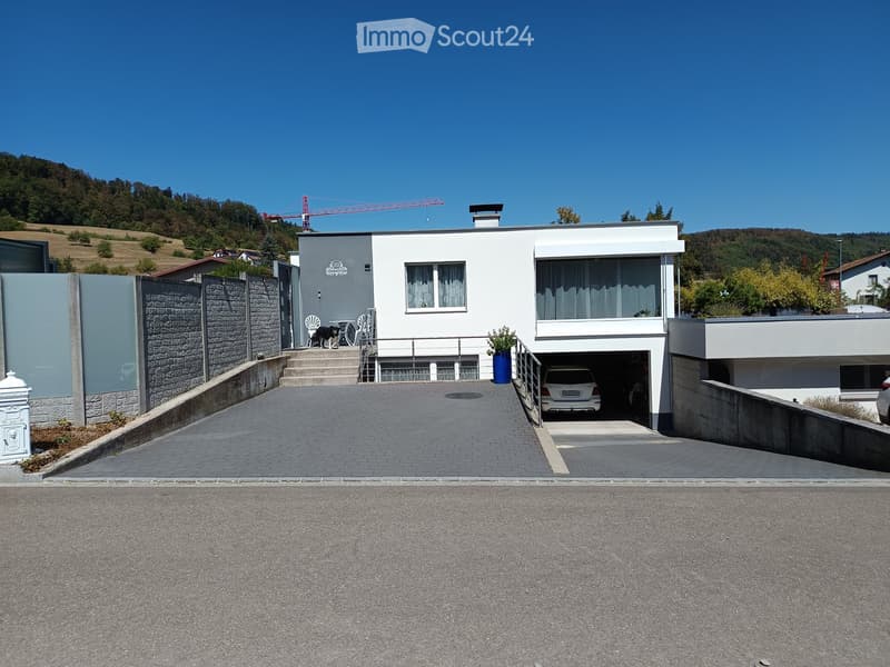 Modern two-story house with white exterior, garage, and outdoor seating area. The house features large windows, a stone wall, and a paved driveway.