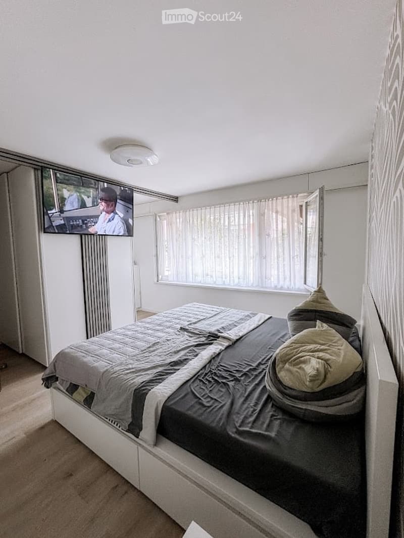Modern bedroom with a large bed, pillows, blanket, mounted television, and a radiator. The room has large windows with sheer curtains and a light-colored carpeted floor.