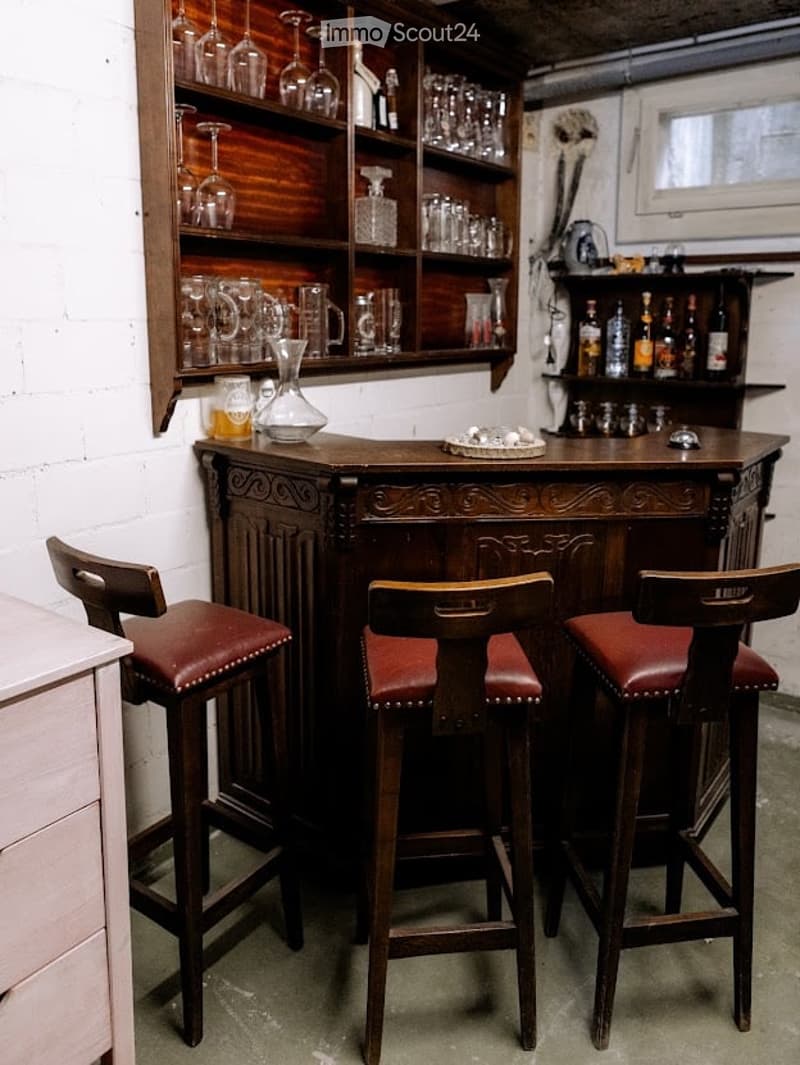 A cozy bar area with a wooden bar counter, three leather-topped bar stools, and shelves filled with glassware and bottles.