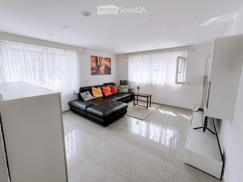 Modern living room with a black leather sectional sofa, colorful throw pillows, a small wooden coffee table, a large screen, and a painting on the wall. The room features large windows with white curtains and recessed ceiling lighting.