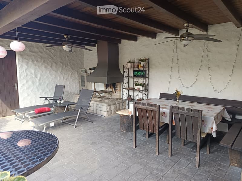 Outdoor patio with stone walls, tiled floor, and a fireplace. Includes a dining table with chairs, lounge chairs, a bench, and a shelf with plants and decorative items.
