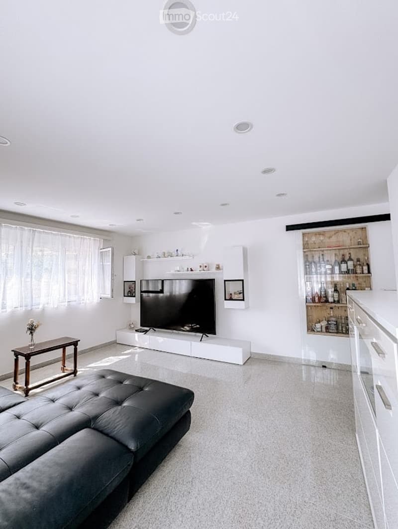 Modern living room with white walls, grey carpet, large windows, a black leather sofa, a wooden coffee table, a flat-screen TV on a white cabinet, and a glass cabinet with bottles and decorative items.