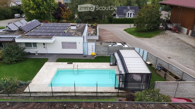 Modern house with a swimming pool in the backyard, surrounded by a fence, with solar panels on the roof and a covered patio area.