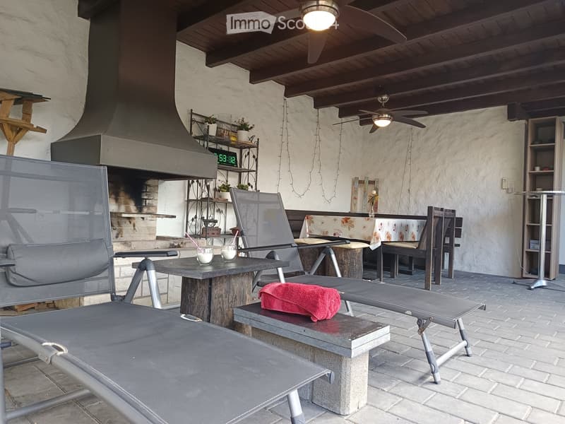 Outdoor patio with a stone table, chairs, a bench, and a small shelf with plants. A ceiling fan is visible, and the area is covered with a wooden ceiling.