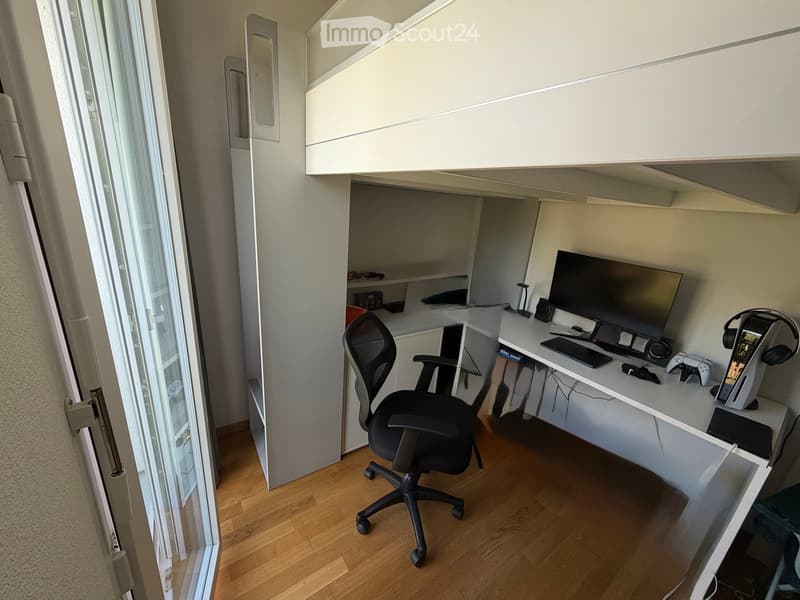 Modern, minimalist workspace with a white desk, ergonomic chair, computer monitor, keyboard, mouse, headphones, and gaming console. The room features light wooden flooring and built-in white cabinetry.
