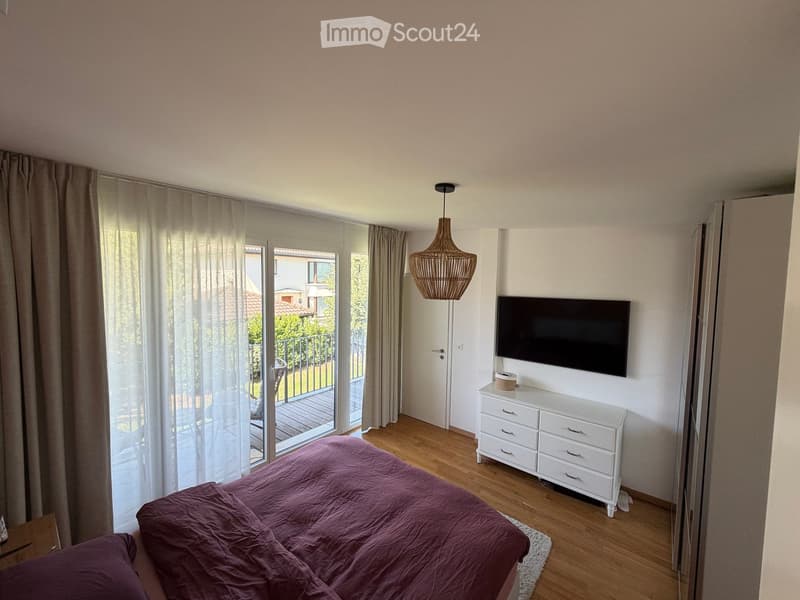 Bright bedroom with wooden flooring, large window with sheer curtains, balcony access, bed with purple bedding, white dresser, television mounted on wall, and a wicker pendant light.