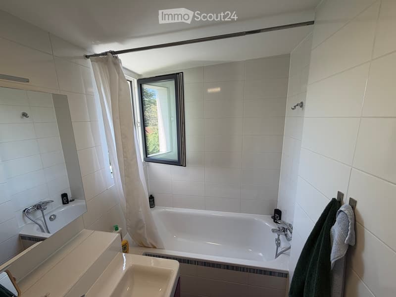 Modern bathroom with a bathtub, sink, mirror, and towel rack. Features a large window with a view of greenery outside.