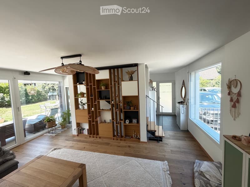 Bright living room with wooden flooring, large windows, a modern bookshelf, a hanging fan, and a cozy seating area with a sofa and coffee table.