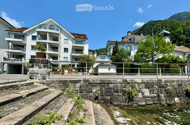 A multi-story white building with balconies and a stone staircase leading down to a small water body, surrounded by greenery and other buildings.