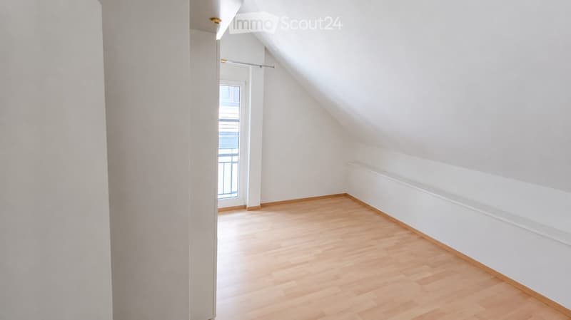 Empty attic room with wooden flooring, white walls, and a window with a view of the outside.