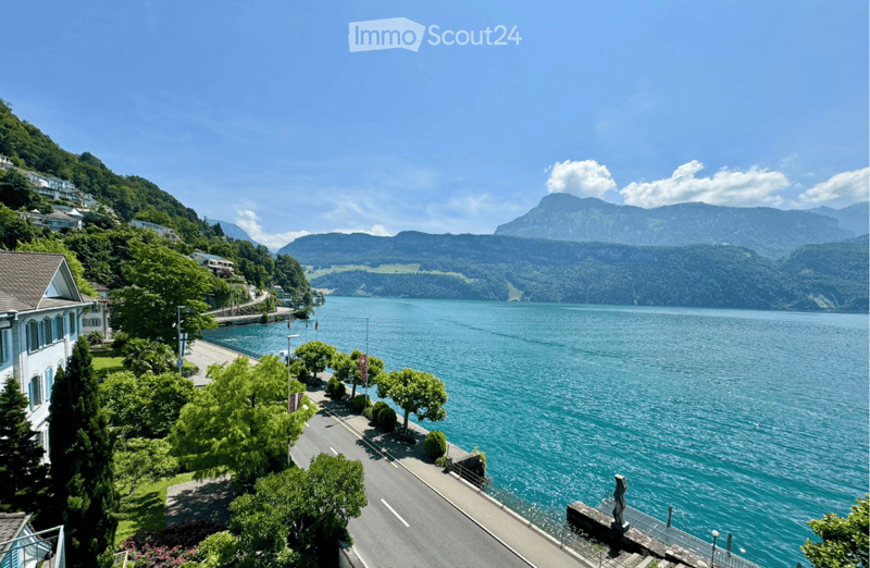 Scenic lakeside view with a clear blue lake, surrounding mountains, and a paved road lined with trees and shrubs.