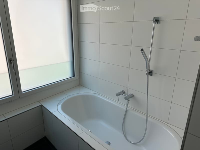 Modern bathroom with a white bathtub, chrome faucet, and handheld showerhead. The walls are tiled in white, and there is a large window with frosted glass on the left side.