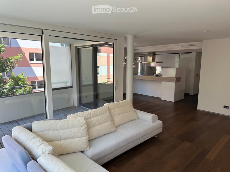 Modern living room with white leather sofa, wooden flooring, large glass sliding doors leading to a balcony, and an open kitchen area with a kitchen island.