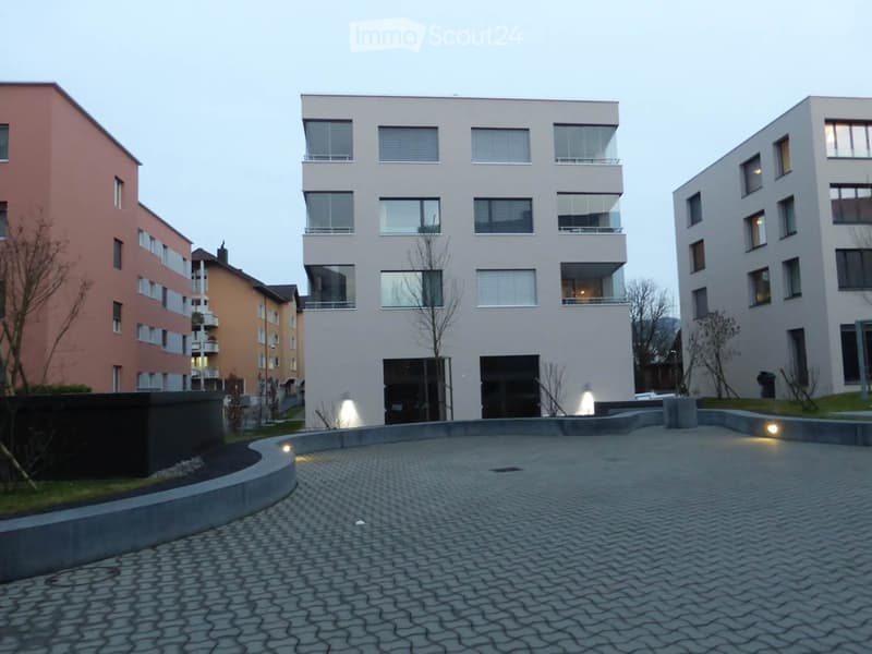 Modern multi-story building with large windows and balconies, surrounded by paved area and low lighting.