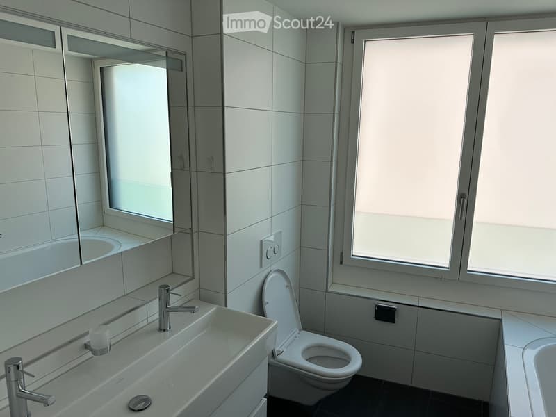 Modern bathroom with white tiles, a large mirror, a sink with a modern faucet, a toilet, and a bathtub. The room is well-lit with large windows.