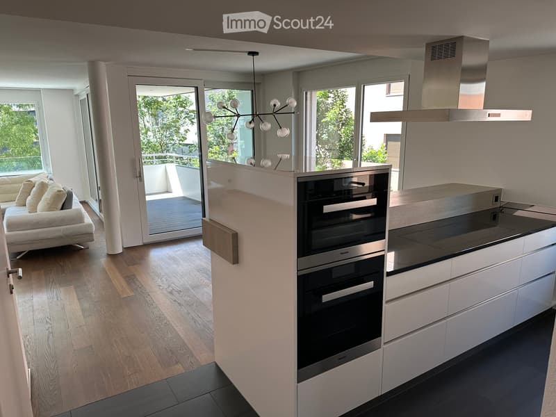 Modern open-plan living and kitchen area with light wood flooring, white walls, and large windows. Features a white sofa, a sleek oven, kitchen cabinets, and a chandelier.