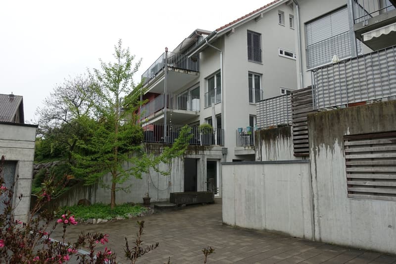 Modern multi-story residential building with balconies, large windows, and a paved courtyard area with greenery and a small tree.