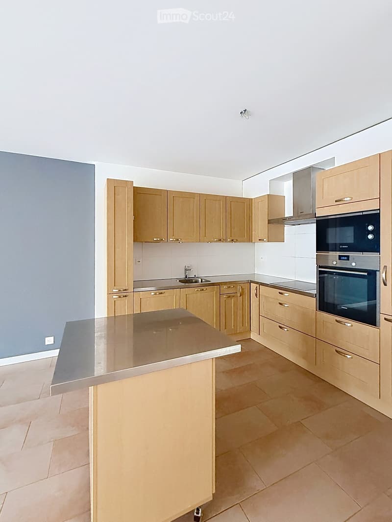 Modern kitchen with light wooden cabinets, stainless steel appliances, and a central island with a glass countertop.