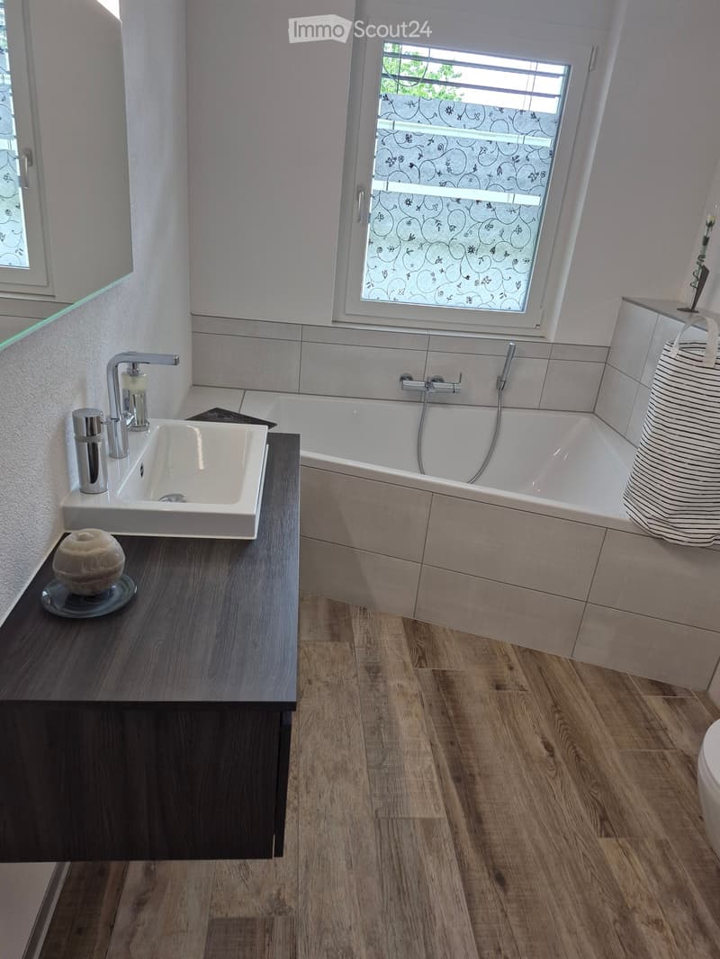 Modern bathroom with a white sink, bathtub, and wooden flooring. Features a large window with decorative glass, a dark wooden vanity, and a handbag on the edge of the tub.