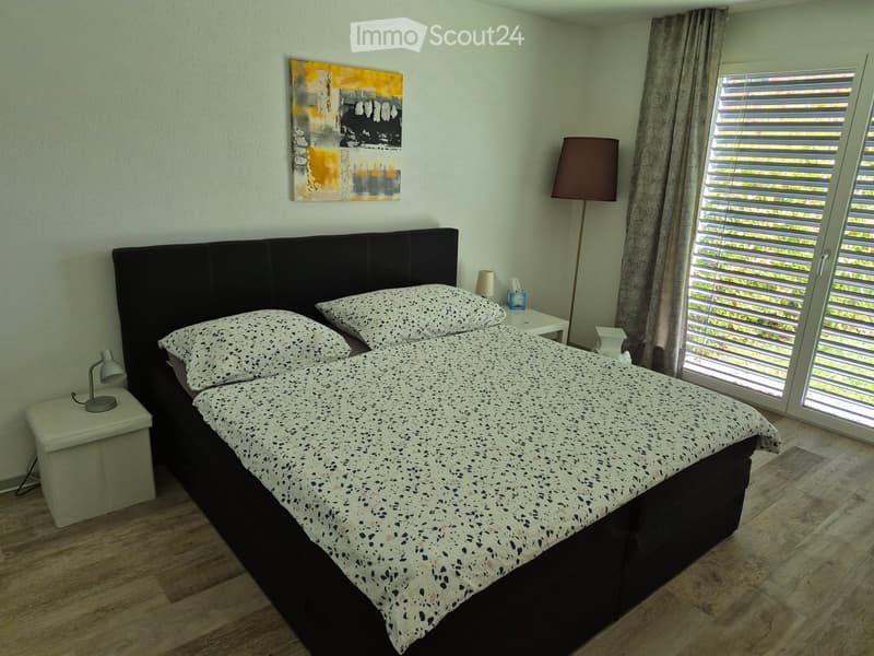Cozy bedroom with a large bed, nightstands, lamps, and a painting on the wall. The room features wooden flooring and large windows with blinds.