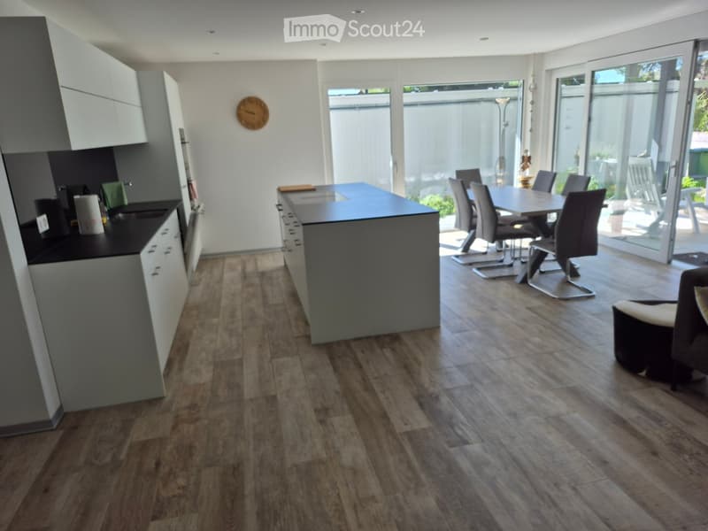 Open-plan kitchen and dining area with modern grey cabinets, a central island, and a dining table with chairs. The space features wooden flooring and large windows leading to an outdoor area.