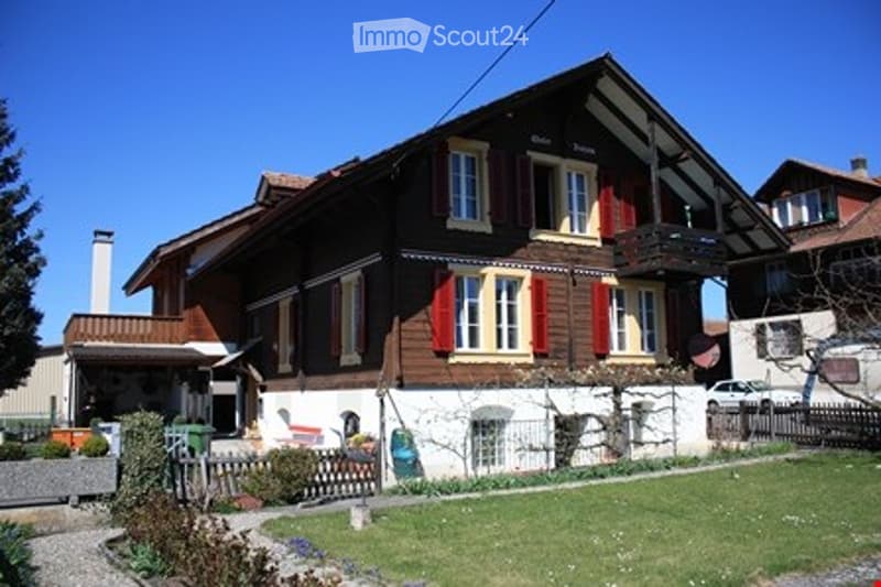 Traditional Swiss house with wooden facade, red shutters, and a small balcony. The house has multiple windows and a stone base, surrounded by a well-maintained garden with a fence and a few plants.