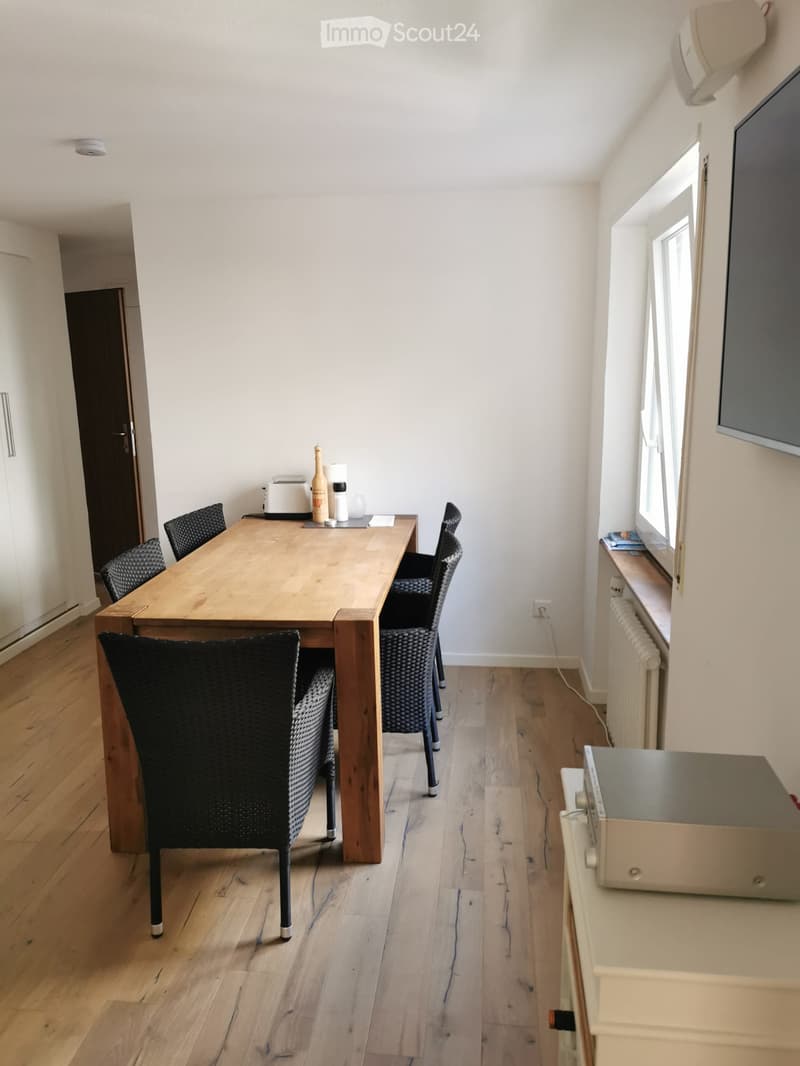 A modern dining room with a wooden table, four black chairs, a small oven, and a screen mounted on the wall. The room features light wooden flooring and white walls.