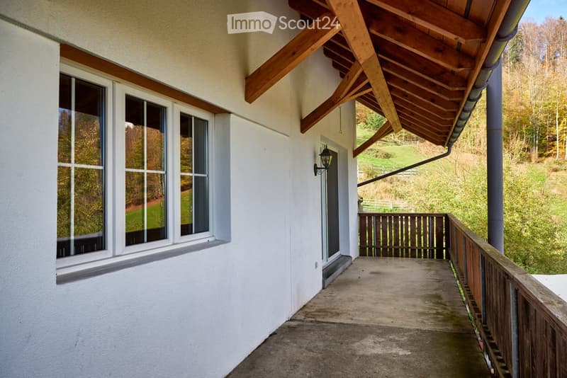 Wooden balcony with a wooden railing, overlooking a green landscape with trees and hills.