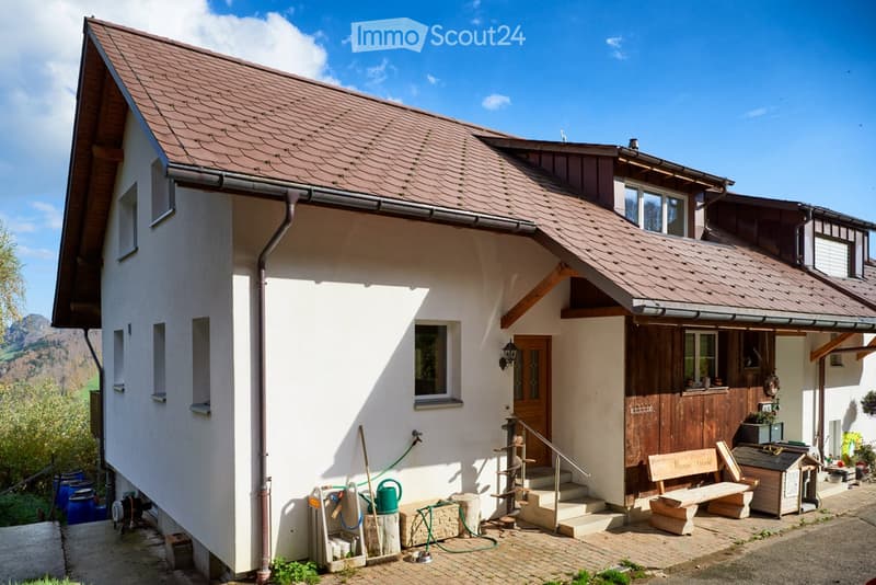 Two-story house with white walls, brown roof, wooden door, and small porch area with bench and tools.