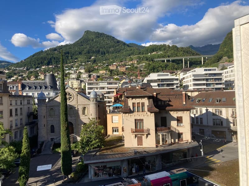 A panoramic view of a town with a mix of modern and traditional buildings, surrounded by green hills and mountains under a partly cloudy sky.