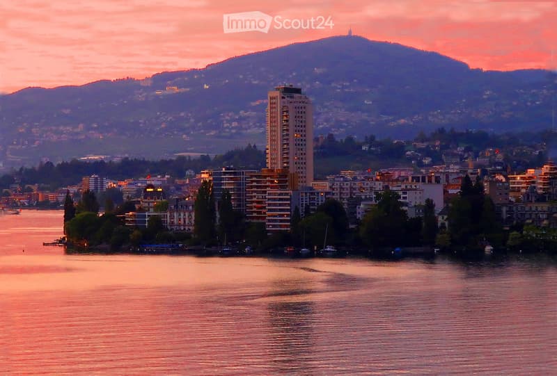 A scenic view of a cityscape at sunset, featuring a body of water in the foreground, with buildings and trees lining the shore, and mountains in the background.
