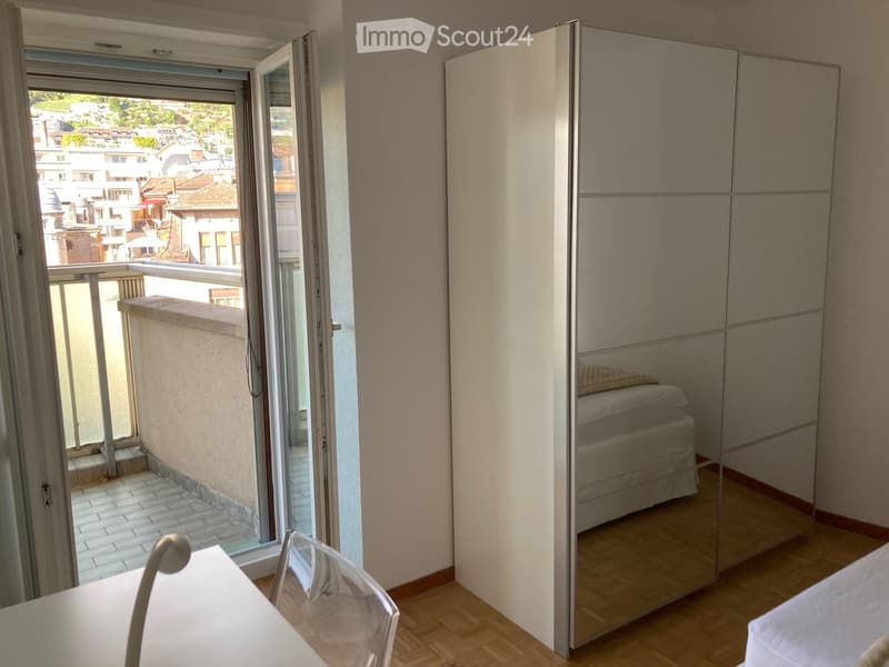 Bedroom with a large mirrored wardrobe, a bed, a small table with a chair, and a balcony overlooking a cityscape.