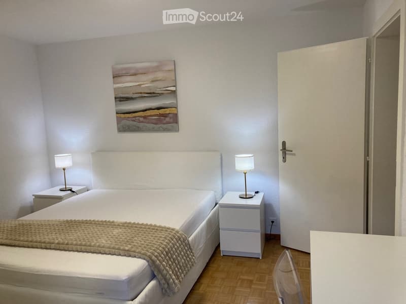 A modern, minimalist bedroom with a white bed, nightstands, table lamps, a chair, and a painting on the wall.