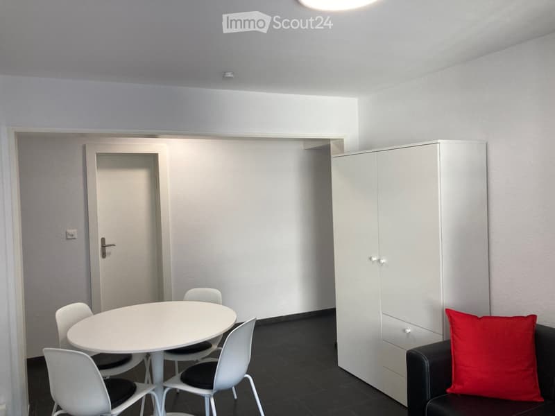 Modern, minimalist room with white walls, black tiled floor, a round white table with four white chairs, a black sofa with a red pillow, and a white cabinet.