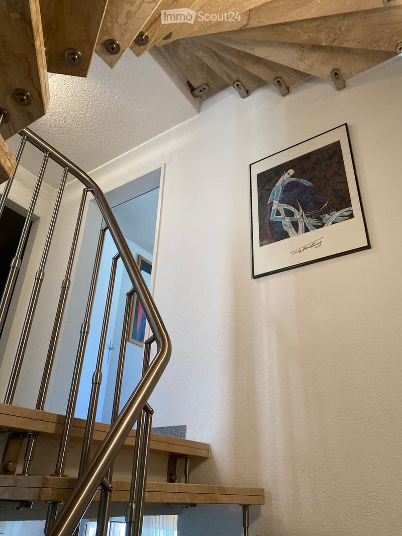 Spiral staircase with wooden steps and metal railing, white walls, framed artwork, and a view of another room through a glass wall.