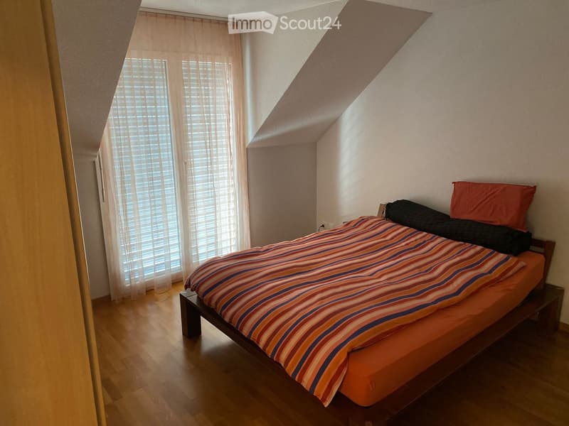 Bedroom with wooden floor, slanted ceiling, large window with blinds and curtains, bed with striped bedding and pillows.