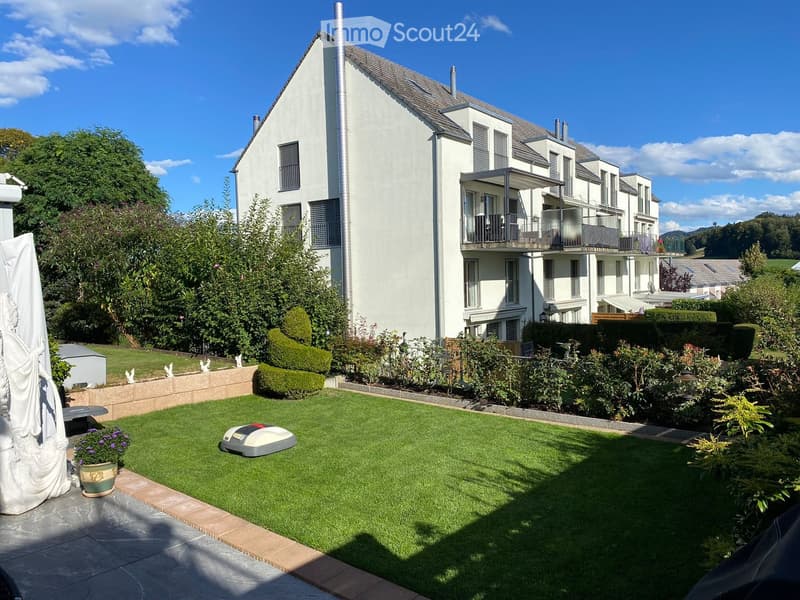 Well-maintained garden with a robotic lawn mower, flower pot, and surrounding greenery, with a modern building in the background.