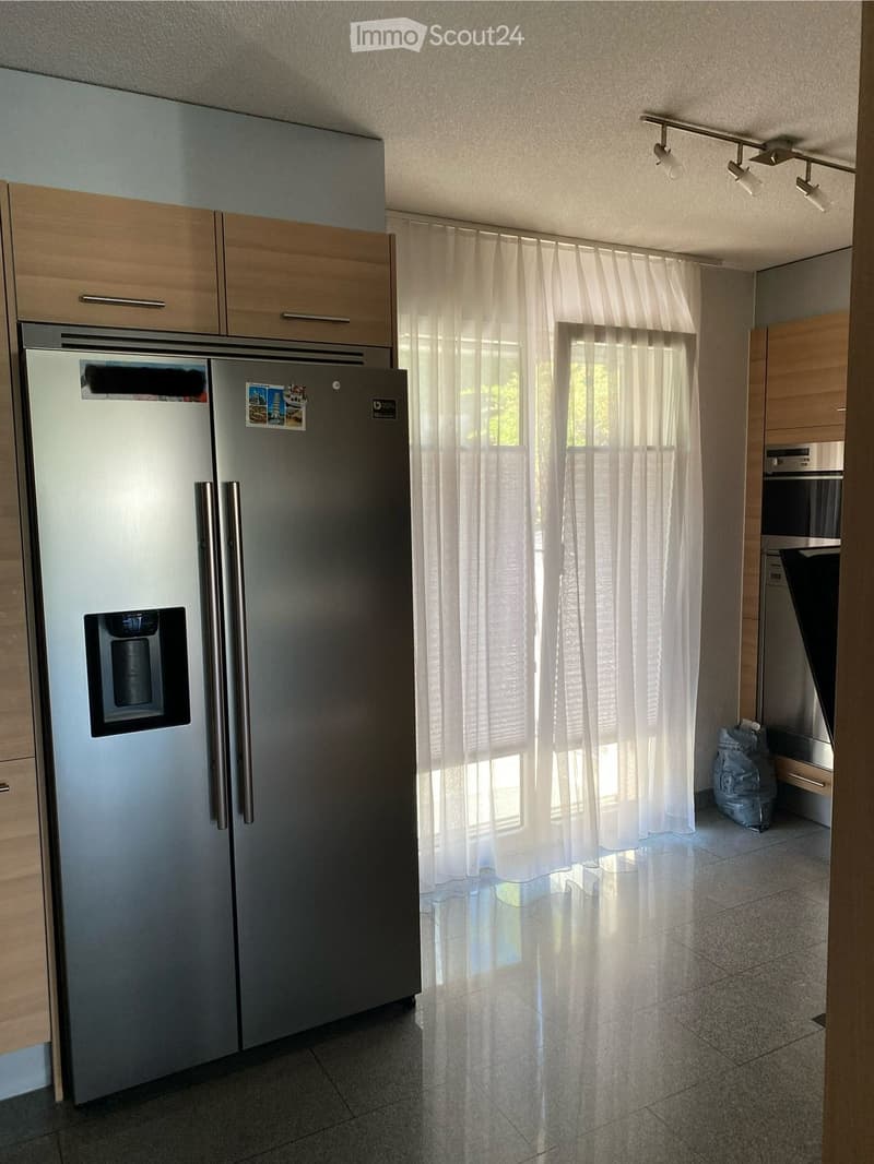 Modern kitchen with stainless steel refrigerator, wooden cabinets, and built-in oven. Light-colored curtains cover the windows, and the floor is tiled.