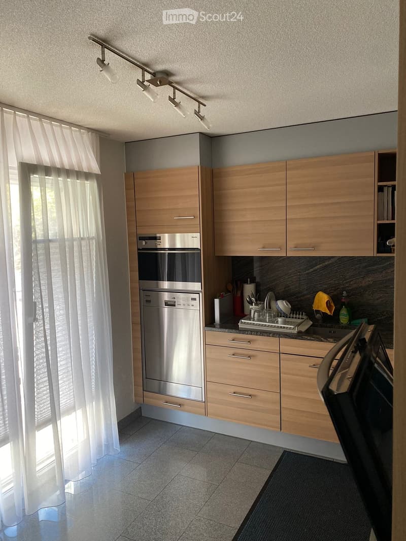 Modern kitchen with light wood cabinets, stainless steel appliances, and a marble countertop. Features an oven, dishwasher, and a sink with a dish rack. Natural light streams in from a window with white curtains.