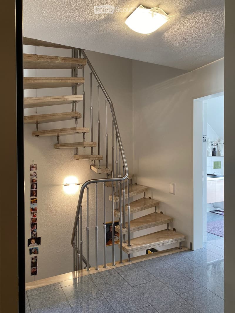 Spiral staircase with wooden steps and metal railing, illuminated by wall-mounted lights, leading to an open area with a glimpse of a bathroom.