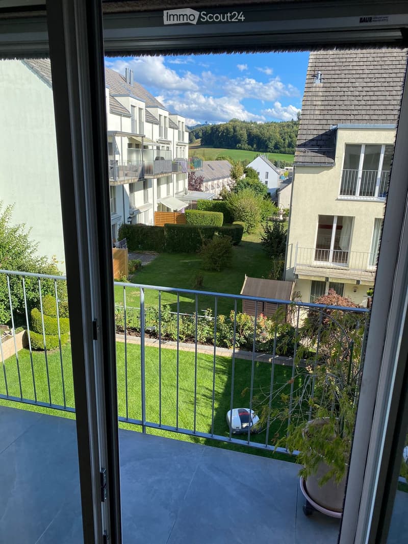 Balcony with metal railing overlooking a well-maintained lawn and garden area, with neighboring houses and greenery visible in the background.